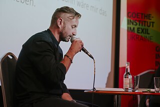 Serhj Zhadan ist von der Seite dargestellt, hält ein Mikrofon in seiner Hand und liest aus seinem Buch "Antenne", das vor ihm liegt, vor.