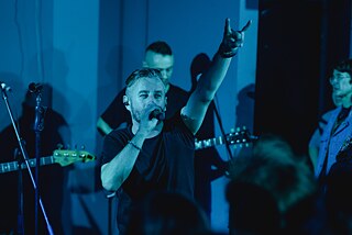 Serhij Zhadan zeigt mit seiner linken Hand die "Metalhand" (englisch "Devil horns"). Hinter ihm ist seine Band "Zhadan I Sobaki" zu sehen. Alle sind schwarz gekleidet. 