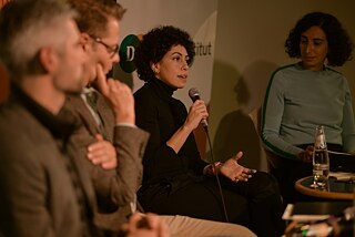 Dima Albitar Kalaji spricht ins Mikrofon, während ihr die neben ihr sitzenden Personen aufmerksam zuhören.