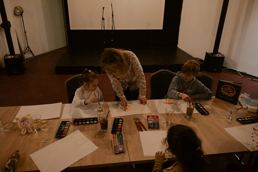 An einem großen Tisch malen drei Kinder mit Wasserfarben. Lana Ra steht in der Mitte und zeigt dem Kind links neben ihr Etwas.