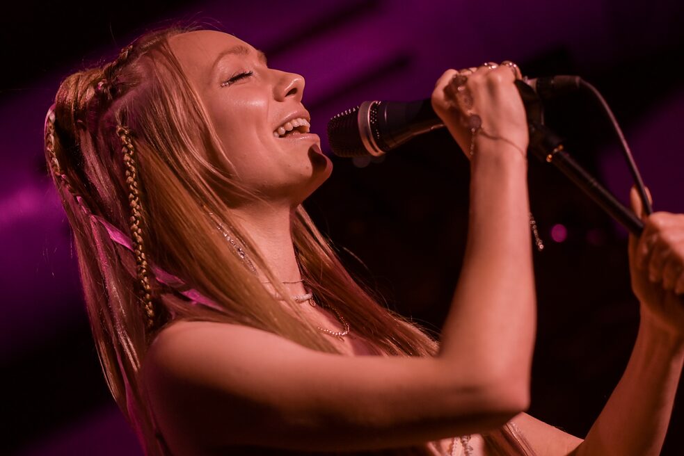 Die junge Sängerin Viktoria ist singend dargestellt. Sie hält das Mikrofon in ihrer Hand fest und die Augen geschlossen.