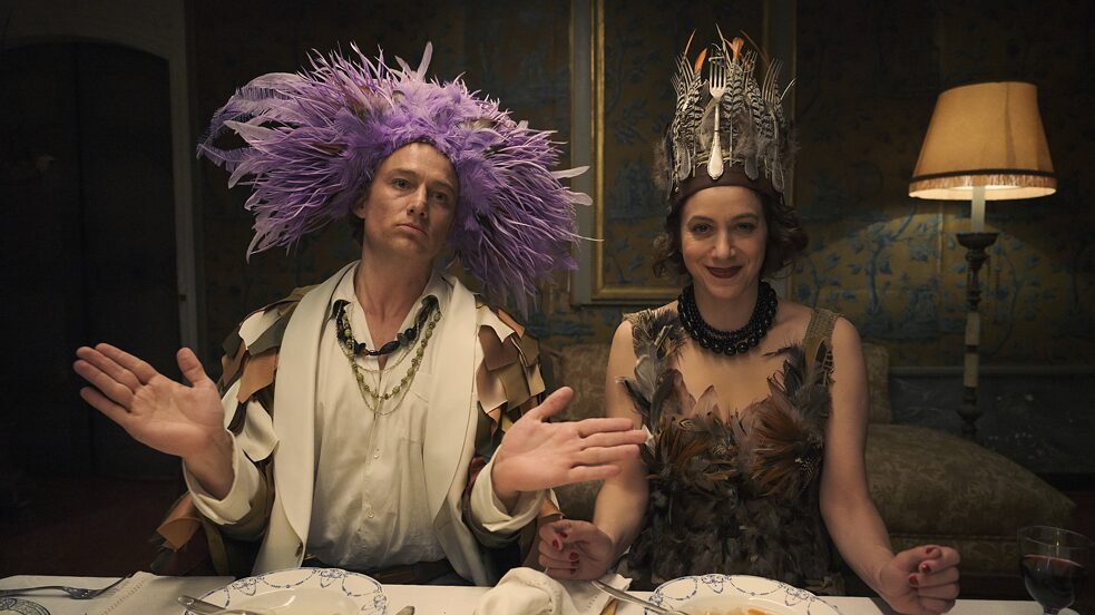 Alexander Fehling, als Künstler Max Ernst, trägt eine Federkrone, während Jodhi May als Kunstförderin Peggy Guggenheim eine Krone mit Gabelzinken trägt. Standbild aus "Transatlantic" von Netflix. 