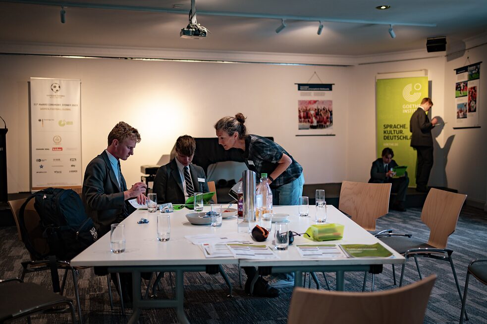 Two students are sitting at a table with teaching materials and a teacher is explaining something to them