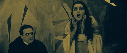 Siyah giyimli bir adamın ağzı açık, şok içinde, bir şeye bakan bir kadına boş gözlerle baktığını gösteren siyah beyaz fotoğraf.