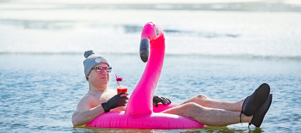 Man with cap, gloves and a cocktail lying on a pink inflatable flamingo surrounded by icebergs