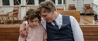 A girl is sitting on a wooden bench with her father, who has her arm wrapped around her shoulder trying to comfort her. 
