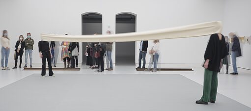 A group of people stand in an exhibition space and watch two performers activating an artwork by Franz Erhard Walther