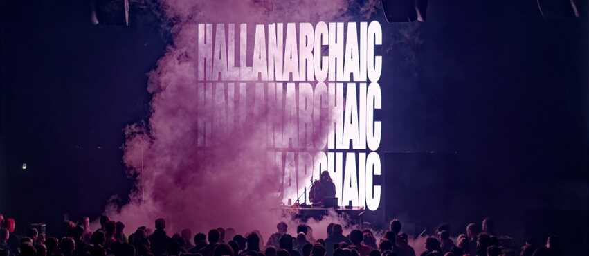 Eine Bühne mit lila Rauch vor der Menge und HALLANARCHAIC auf einem Bildschirm hinter dem DJ