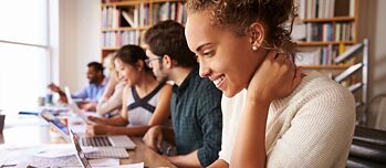 Um grupo de alunos está sentado na biblioteca fazendo um trabalho em grupo. Em primeiro plano está uma jovem com um laptop, ela está sorrindo. A foto é da série "MODERN LIFE" (Vida Moderna).