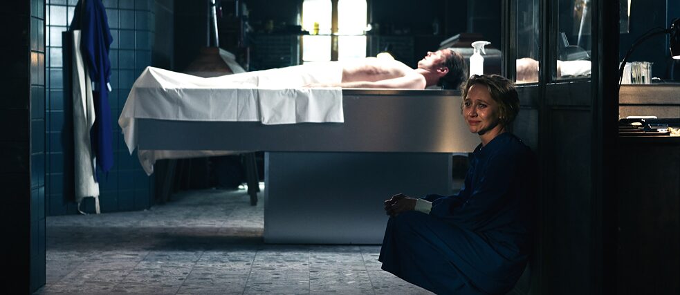 Blum (Anna Maria Mühe) sits crying in the funeral. In the background her husband Mark Thaler (Maximilian Kraus) is lying on the table.