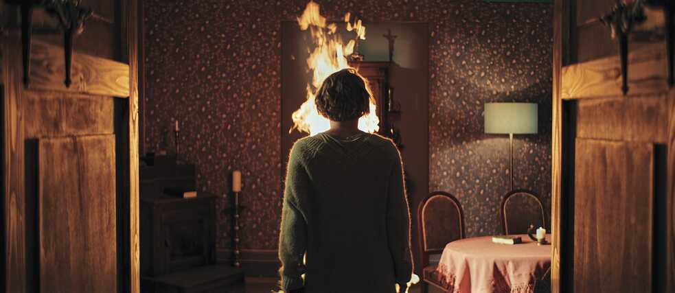 Pastor Jaunig (Simon Schwarz) is tied to a chair and is on fire. Blum (Anna Maria Mühe) watches.