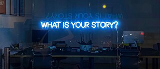Photo is showing Neon lights in a window, spelling out „what is your story“.