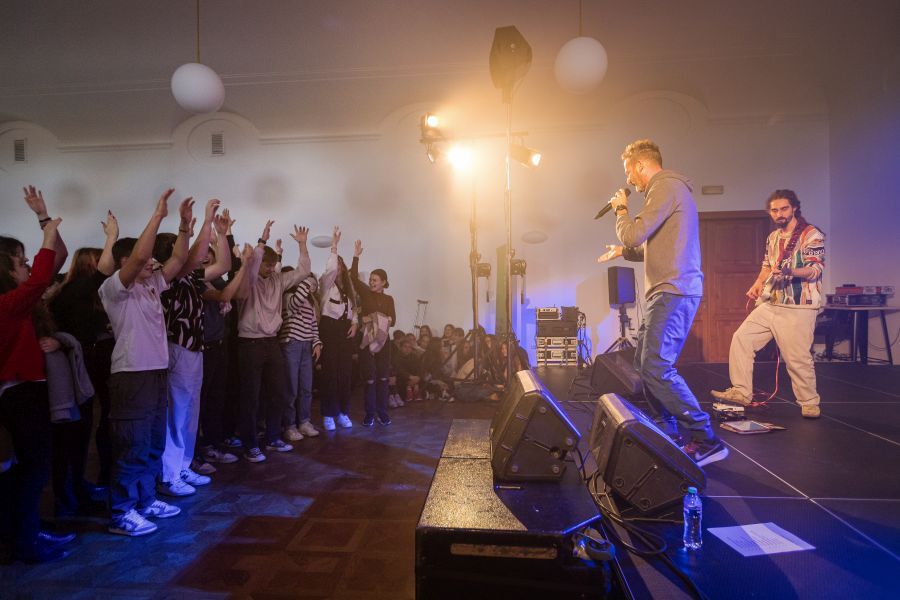 Kapela stojí na pódiu vpravo, zatímco diváci zvedají ruce do vzduchu vlevo.