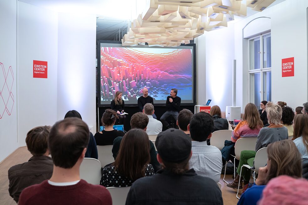 Approx. 70 people watching 3 people speak in front of a screen showing a colourful image. The scene is shown from the audiences perspective in a room at Einstein Center Digital Future, Berlin.