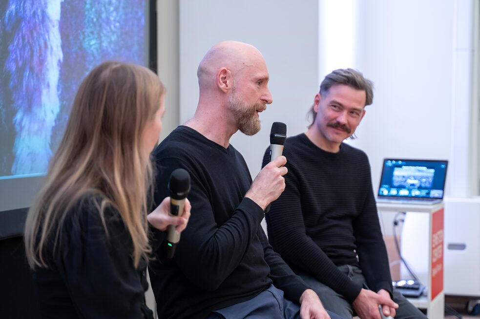 Three people are shown in conversation. The person in the middle, Roman Lipski is holding a mic and talks while Roisin Kiberd to his right and Tim Schröder to his left are looking at him and listening to him.