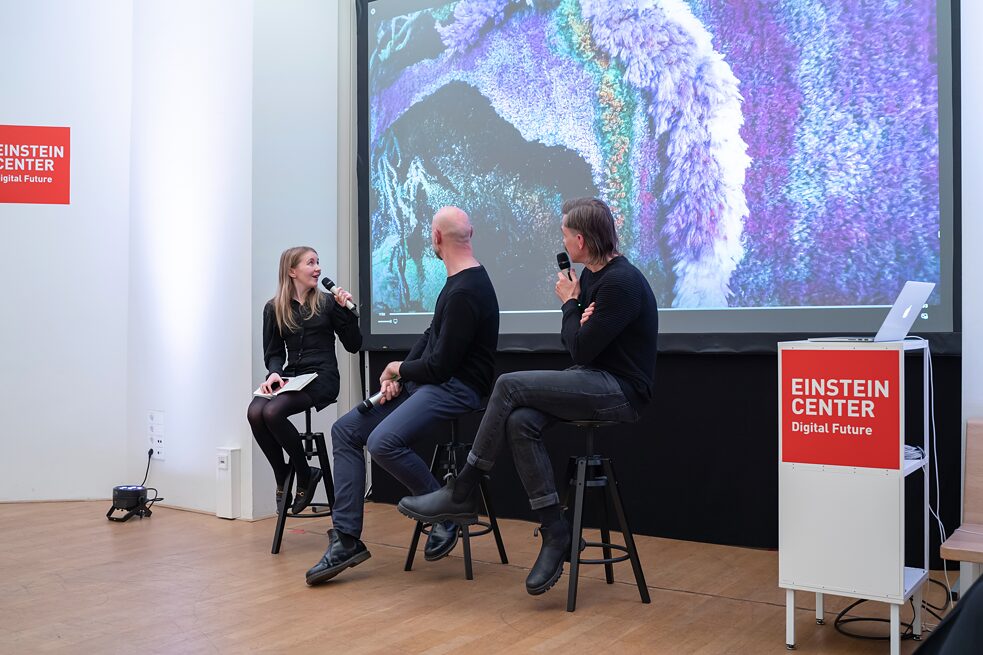 Roisin Kiberd, Roman Lipski and Tim Schröder are sitting on stools in front of a screen holding microphones loosely in their hands. Tim is speaking into his mic while Roisin is looking at him and Roman is looking into the distance.