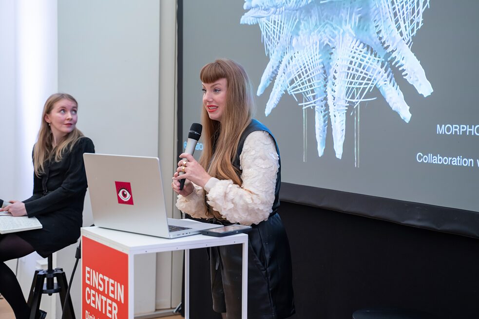 Artist Amy Karle is standing behind a lecturn with her laptop on it. She is standing in front of a screen and speaking into a mic. In the backgraound, Roisin Kiberd is sitting on a stool and is looking over her shoulder at the screen.