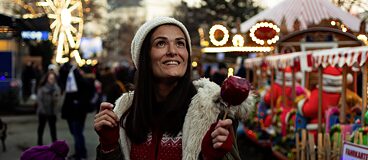 Gabriela in Deutschland. Sie geht Abends auf einem Weihnachtsmarkt spazieren. Sie hält einen roten, kandierten Apfel in der Hand.
