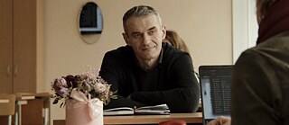 Photo is showing the middle-aged man Slava sits at his desk at school. 