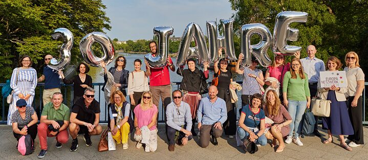 Alumni-Gruppe während des Alumnitreffens 2023 in Berlin steht auf einer Brücke und hält silberne Lutballons hoch, die den Schriftzug 30 Jahre ergeben