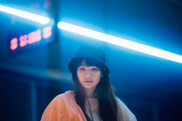 Die Künstlerin Edy Fung schaut direkt in die Kamera. Sie trägt einen schwarzen Hut und steht vor einem dunklen, verschwommenen Hintergrund. Im Hintergrund sind ein rotes Leuchtschild und eine bläulich-weiße Neonleuchte zu sehen. 