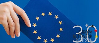 Eine Hand hält einen Wahlschein mit der Europa-Fahne über eine Wahlurne zur Europawahl 2024
