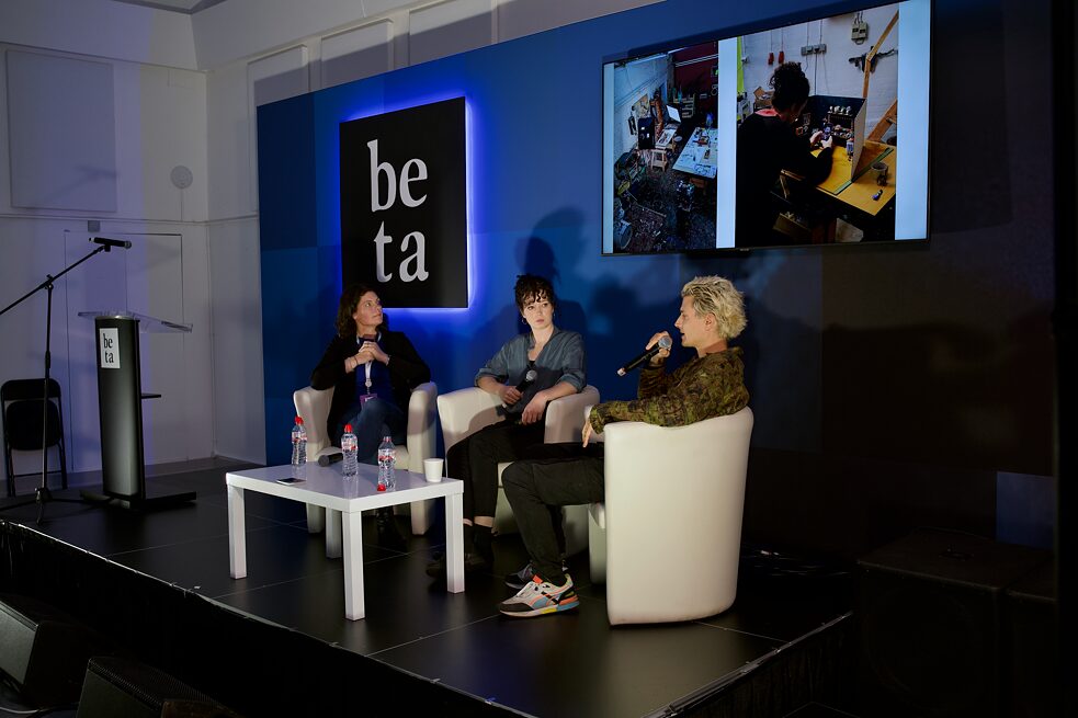 3 people are pictured sitting in white chairs on a stage with a small white table in front of them. The woman on the left is looking up at a screen on the wall behind them which shows images of the artists' studios. the woman at the centre is looking at the man on the right who is speaking into a micropone.