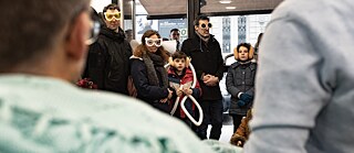 Im Vordergrund die Rücken von zwei Männern. Im Hintergrund mehrere Menschen mit skurrilen Brillen, Ohrenschützern und Schläuchen um den Hals