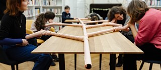 Mehrere Menschen sitzen an einem Tisch und halten ihre Ohren an einen Schlauch, der auf dem Tisch liegt