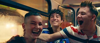 Drei lachende Teenager sitzen in öffentlichen Verkehrsmitteln und schauen schelmisch.