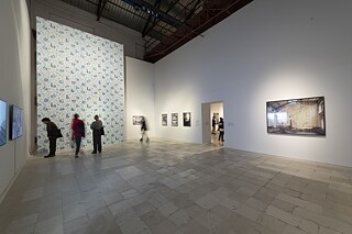 Ausstellungsansicht mit einer großen Tapeteninstallation und gerahmten Fotografien an den Wänden. Die Besucher bewegen sich in der Halle.