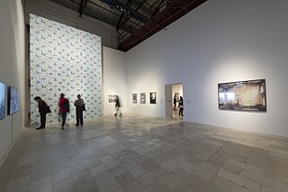 Exhibition view with a big wallpaper installation and framed photographs on the walls. Visitors are moving around in the hall.