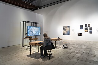 A girl is sitting on a stool at a wooden desk and listens to something with headphones. In the room is another desk and stool, a metal and wood installation with a video screen and photographs on the wall.