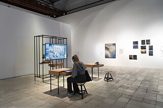 Ein Mädchen sitzt auf einem Hocker an einem Holzschreibtisch und hört mit Kopfhörern etwas. Im Raum stehen ein weiterer Schreibtisch und ein Hocker, eine Installation aus Metall und Holz mit einem Videobildschirm und Fotos an der Wand.