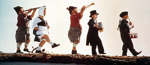Fermo immagine tratta dal film “Die Blechtrommel”: cinque persone suonano degli strumenti musicali mentre camminano su un tetto