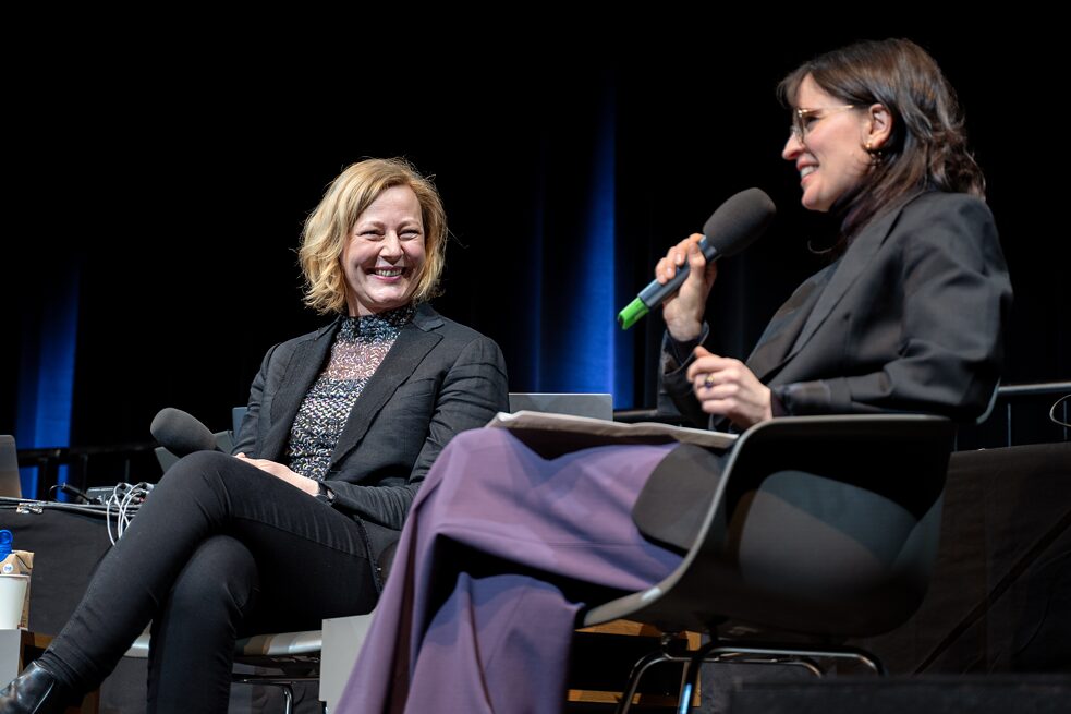 Gesche Joost and Clara Herrmann are sitting on black chairs on a stage. They hold microphones and smile. Clara is speaking.
