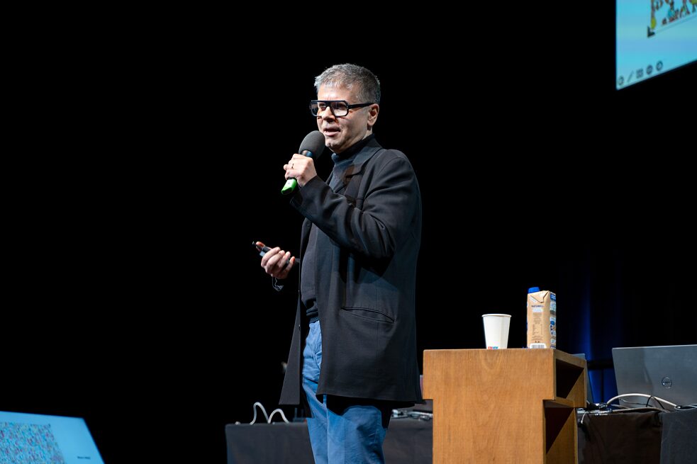 Eduardo Miranda is standing on a stage speaking into a microphone. Beside him is s small, brown table and a screen is visible in the top right corner of the image.