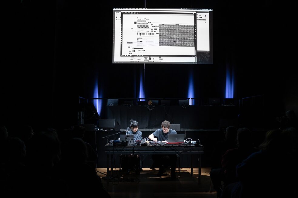Edy Fung and Muiredach O’Riain are sitting behind a desk on a stage. They operate computers. On the wall behind them is a screen that shows their artwork. 