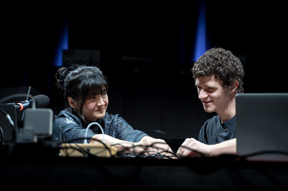 Edy Fung and Muiredach O’Riain are sitting on a stage behind a desk. They are operating computers and pushing buttons, smiling at each other 