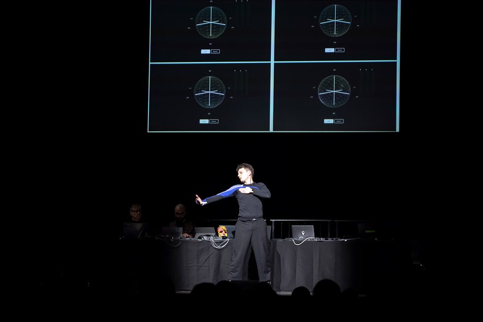 A person is standing on a stage in front of a desk. On the wall behind them is a screen. The person is looking at their outstretched right hand. The person's left hand is placed on their chest.