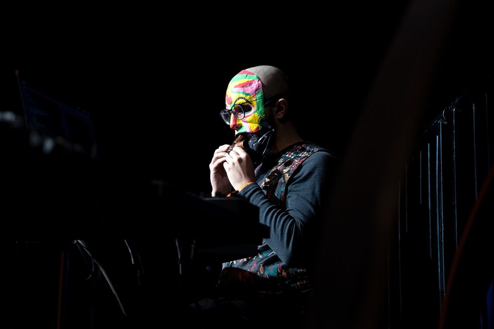 A person wearing a brightly coloured mask is sitting behind a desk on a stage. They are blowing air into a wooden instrument.