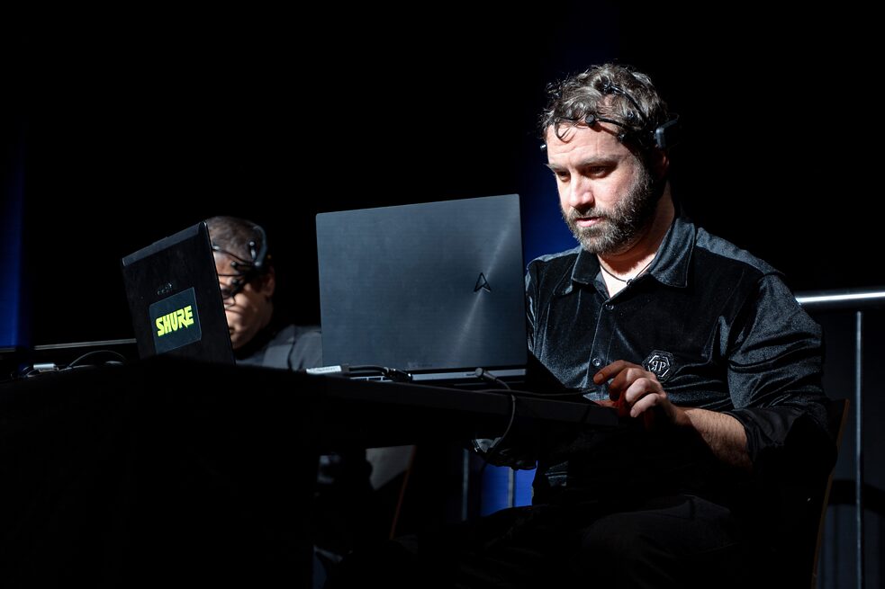 Two people are sitting behind a desk on a stage. On the desk are computers. The person in the foreground is wearing a black device on their head that transmits data from their brain to the computer in front of them.