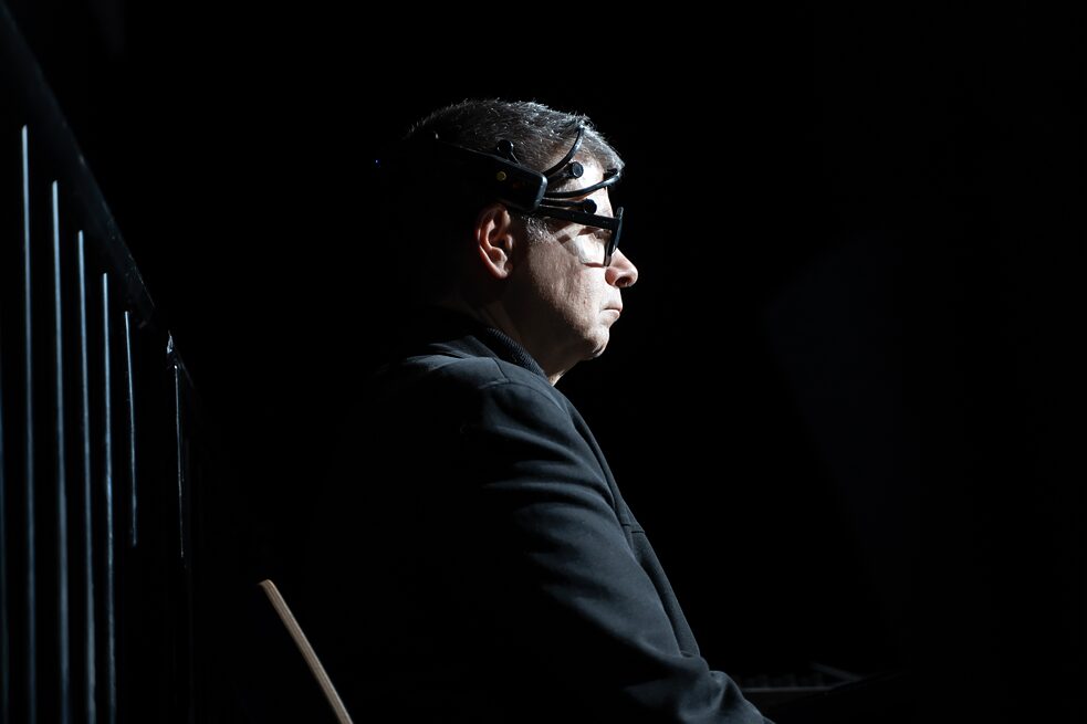 Eduardo Miranda is sitting on a chair on a stage. He is staring at something in front of him and wearing a black device on his head.