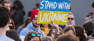 Photo is showing a demonstration with a woman holding up a sign that says “Stand with Ukraine”.