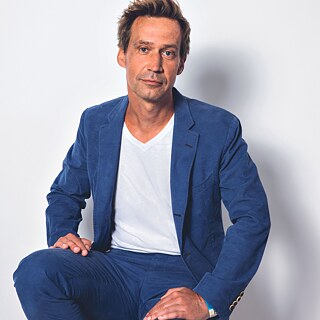 Der Autor Volker Weidermann trägt auf dem Bild einen blauen Anzug und sitzt entspannt auf einem Hocker. Sein Blick ist in die Kamera gerichtet.