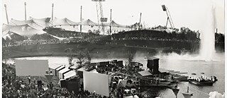 La photo montre la vue depuis le versant de la montagne olympique sur la presqu'île de Buden lors des Jeux olympiques de 1972 à Munich. Un grand nombre de spectateurs/-trices observent les différents événements sportifs.
