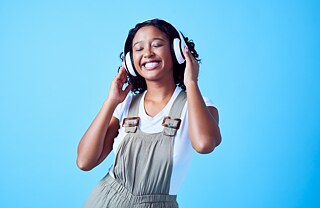 A laughing young woman with closed eyes and white headphones listens to a podcast music, she is in front of a blue background. 