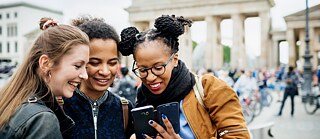 Drei junge Frauen stehen vor dem Brandenburger Tor in Berlin und betrachten lachend die Fotos auf einem Smartphone.  © © Goethe-Institut Living and Working In Germany Amif Events