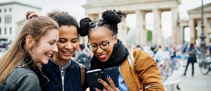 Drei junge Frauen stehen vor dem Brandenburger Tor in Berlin und betrachten lachend die Fotos auf einem Smartphone. 