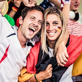 Ein Mann in Fan - Shirt und eine Frau mit um die Schultern gelegtem Deutschland Trikot liegen sich in den Armen und jubeln.
